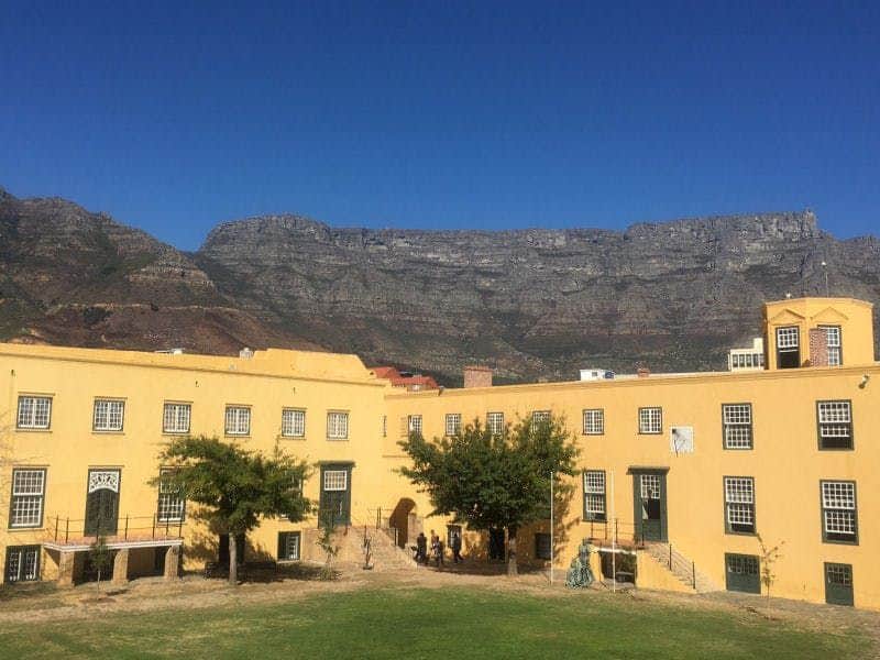 A yellow building with green doors and windows, trees in front, and a mountain range in the background under a clear blue sky.