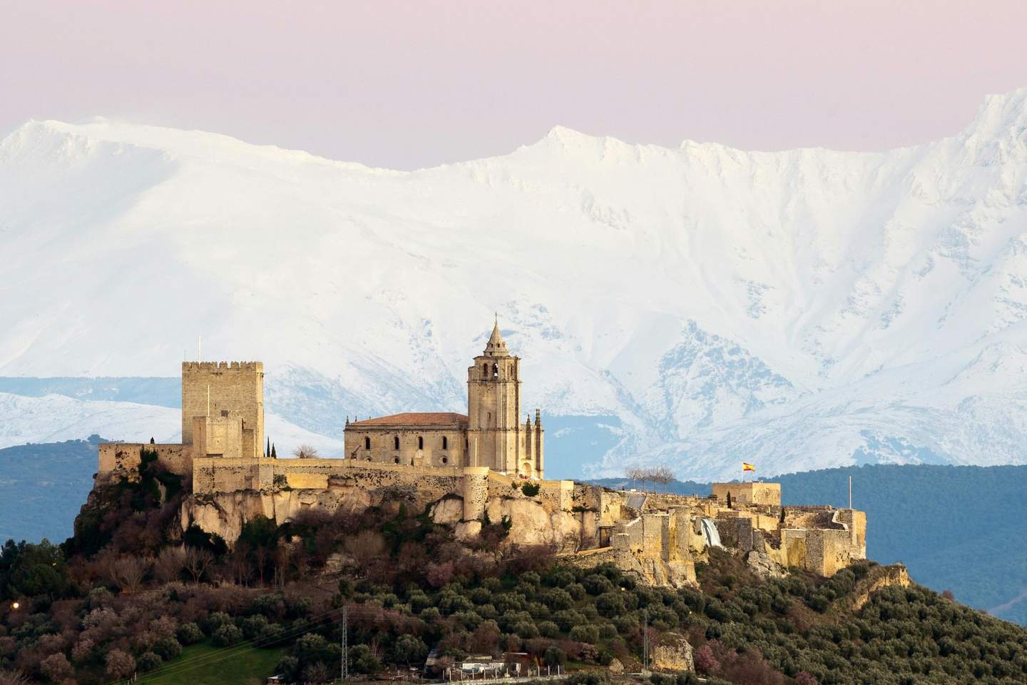 Ancient castle and church on a rocky hilltop with snow-covered mountains in the background, under a pastel pink sky.