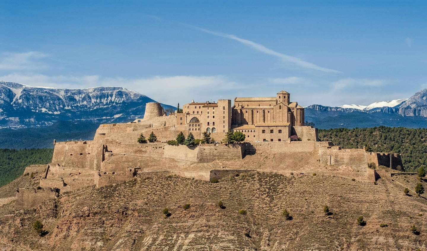 A large stone fortress atop a mountain, with snow-capped peaks in the background and a blue sky above.
