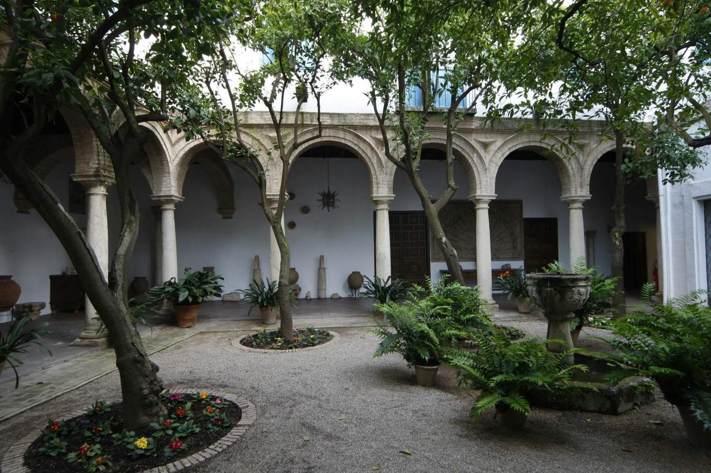 A courtyard with arches, trees, a stone fountain, potted plants, and flowers.