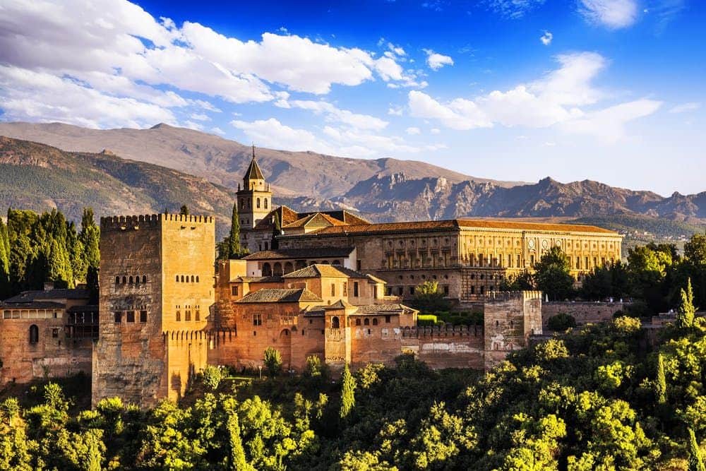 A large, historic fortress with towers and ornate buildings sits amid lush greenery, with mountains and a blue sky in the background.