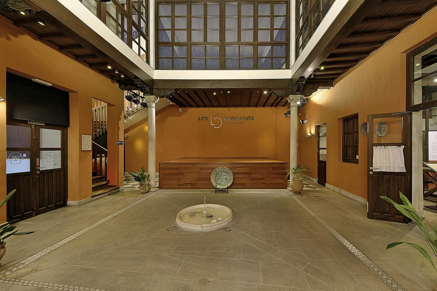 Palacio de los Olvidados Indoor courtyard with orange walls, central circular fountain, wooden doors, and potted plants. "Los Olvidados" sign on wall.