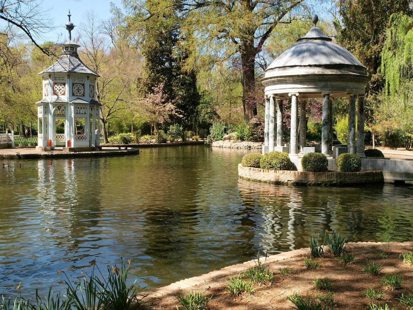 Park scene with a pond, on the left a white gazebo, on the right a round pavilion with columns, surrounded by trees and greenery.