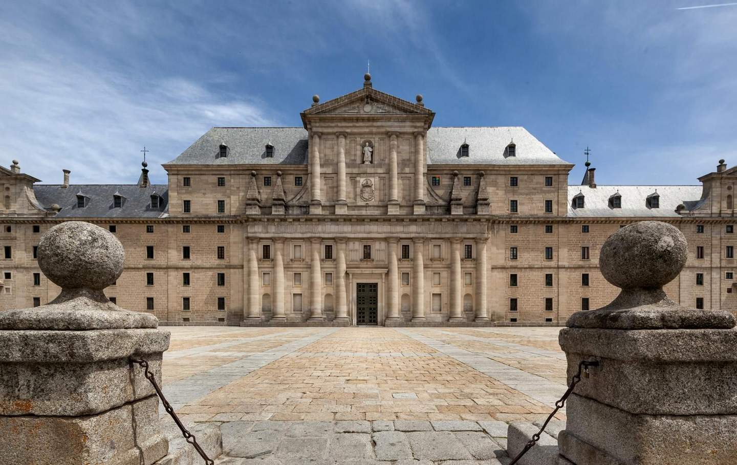 Monastero di San Lorenzo dell'Escorial Un edificio storico con una facciata simmetrica, colonne e una statua centrale, incorniciata da due pilastri di pietra, sotto un cielo azzurro.