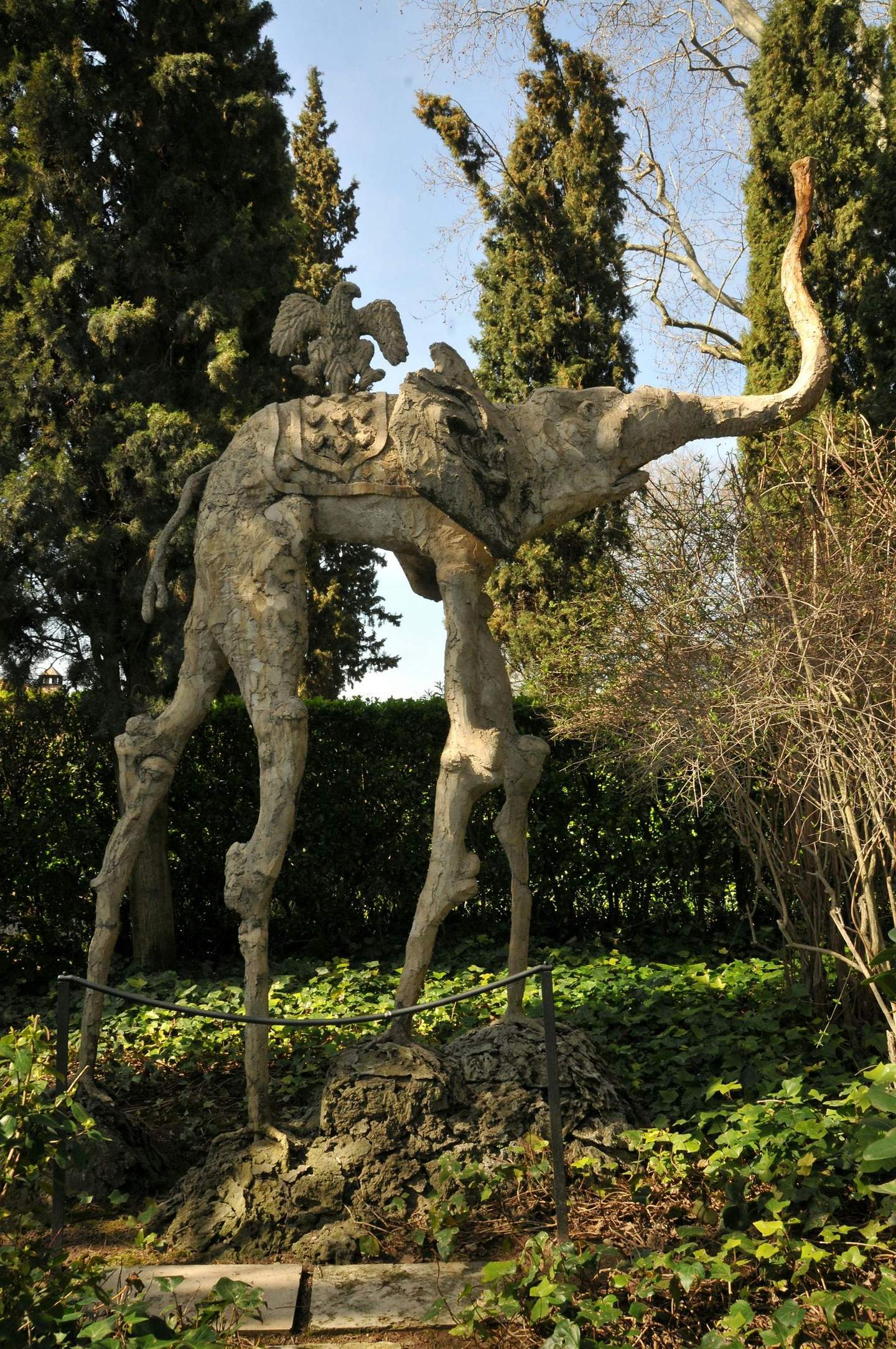 A surreal sculpture of a long-legged elephant with an eagle on its back, surrounded by trees and greenery.
