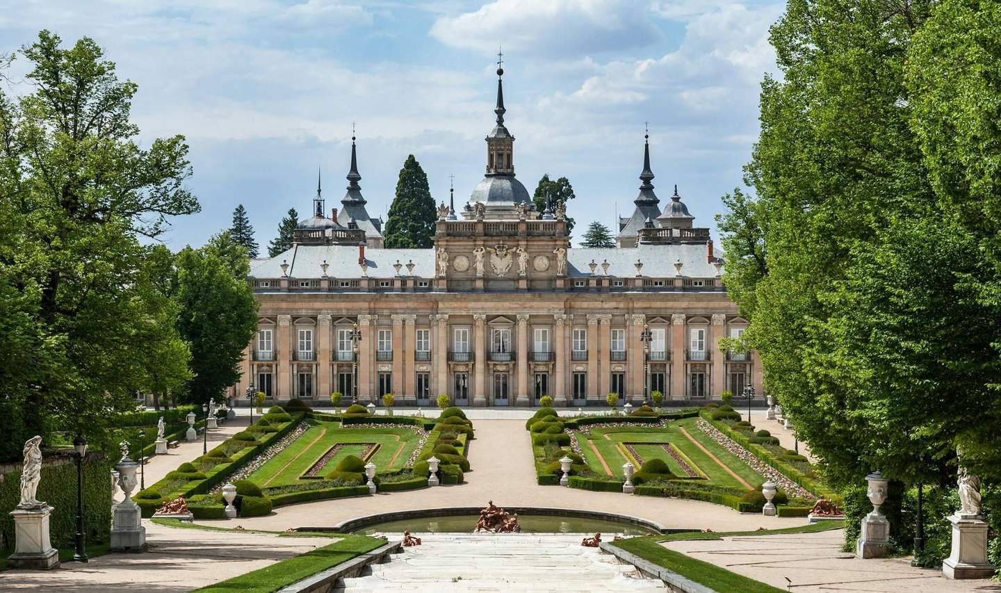 Ornate palace facade with statues, manicured gardens, and a central walkway, flanked by trees and cloudy sky above.
