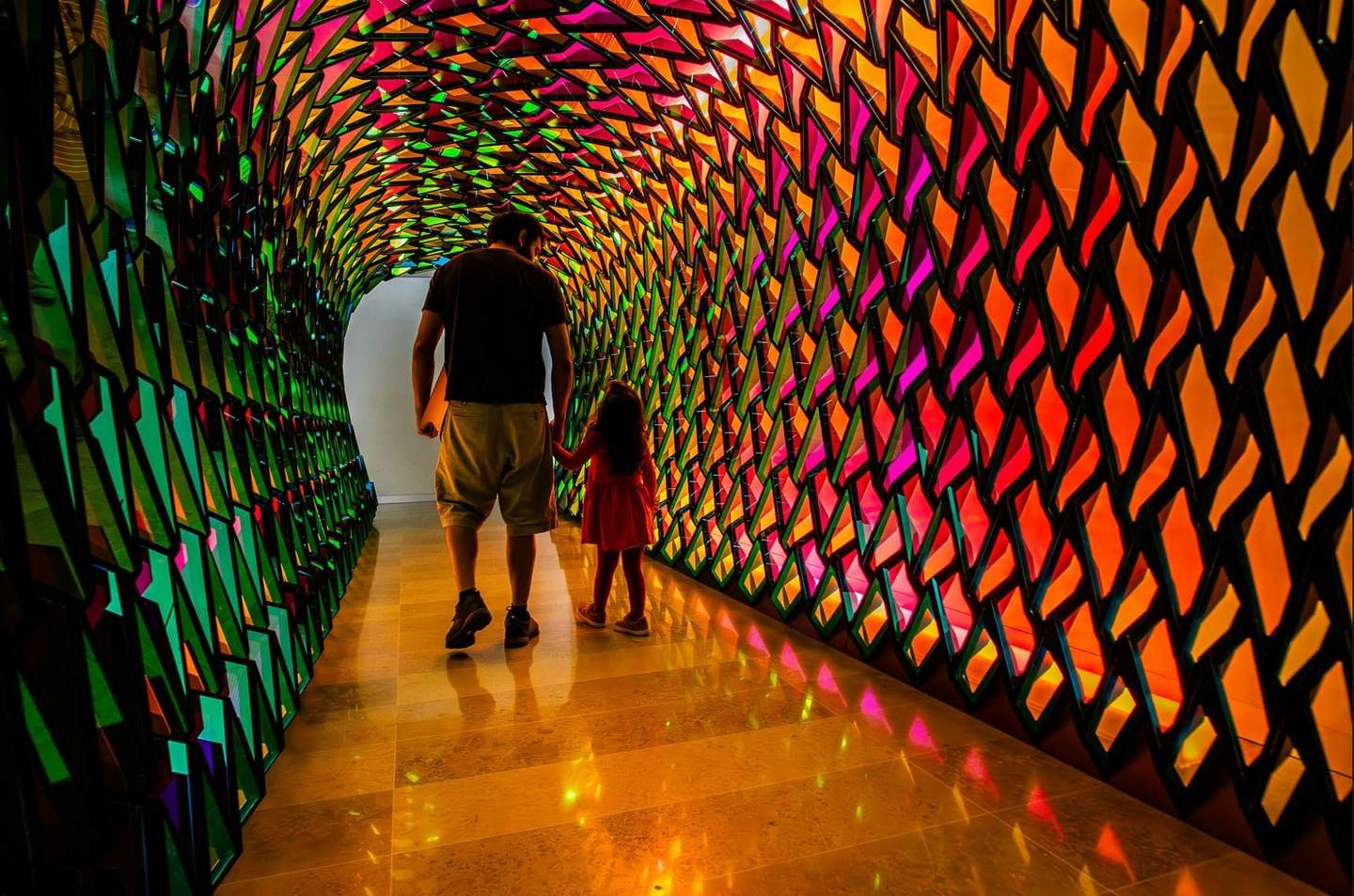 A man and a child walk through a colorful, geometrically patterned tunnel with vibrant lights reflecting on the floor.