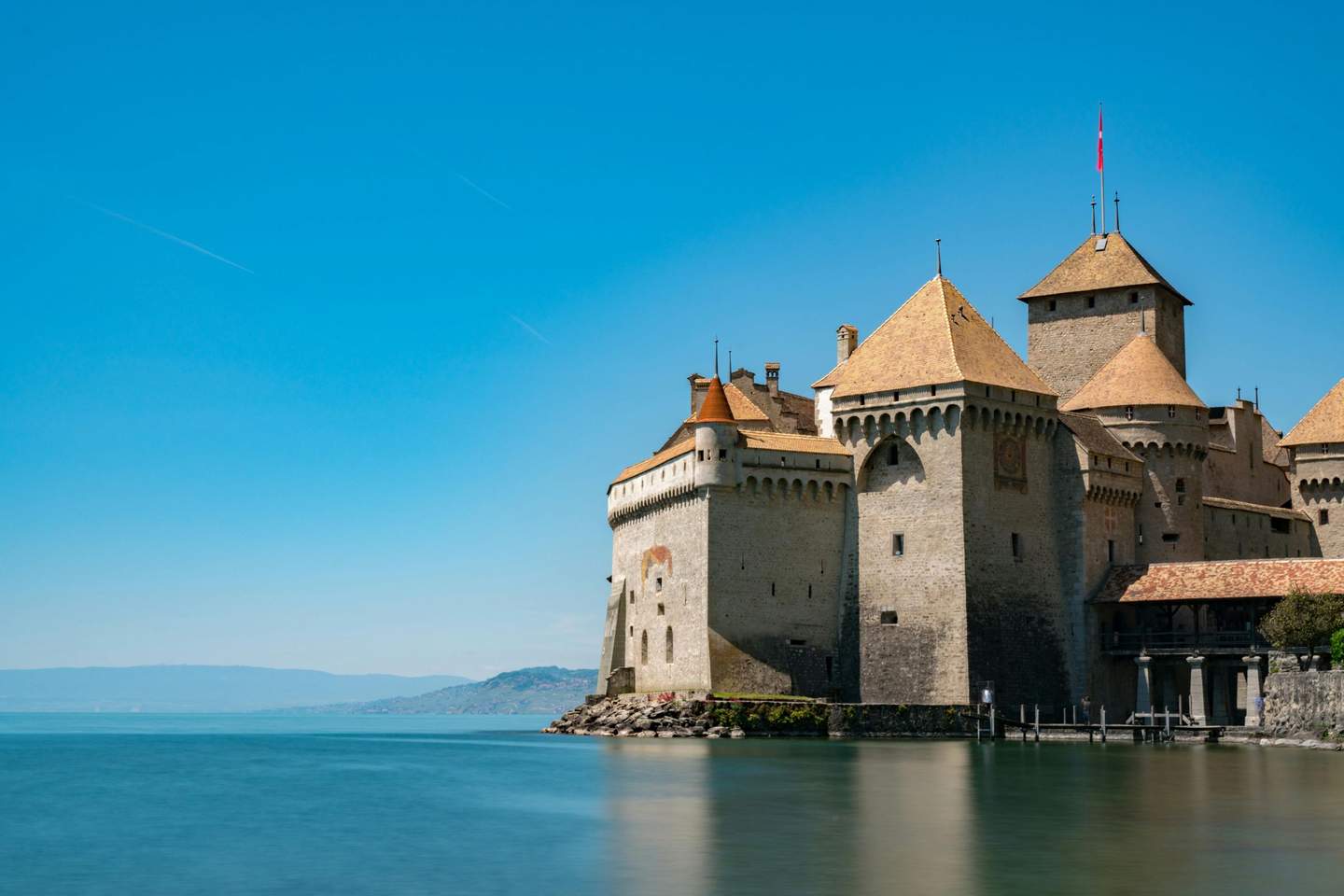 A stone castle with orange-tiled roofs sits by a calm lake, with mountains in the background under a clear blue sky.
