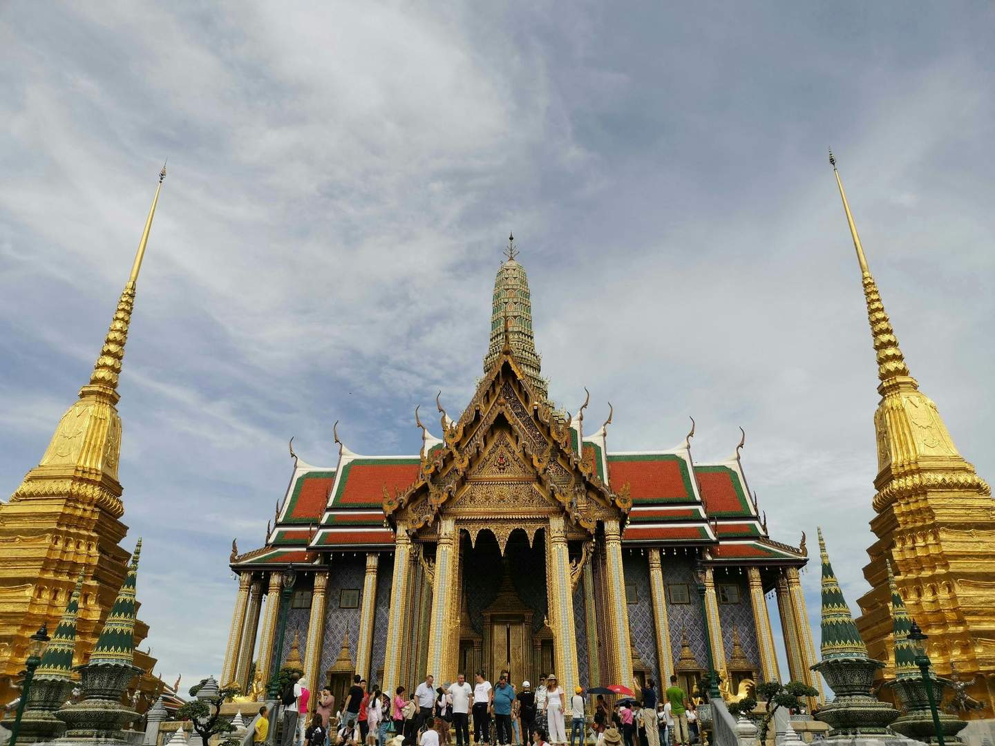 Tourists visit an ornate temple with golden spires, intricate details, and colorful rooftops under a partly cloudy sky.