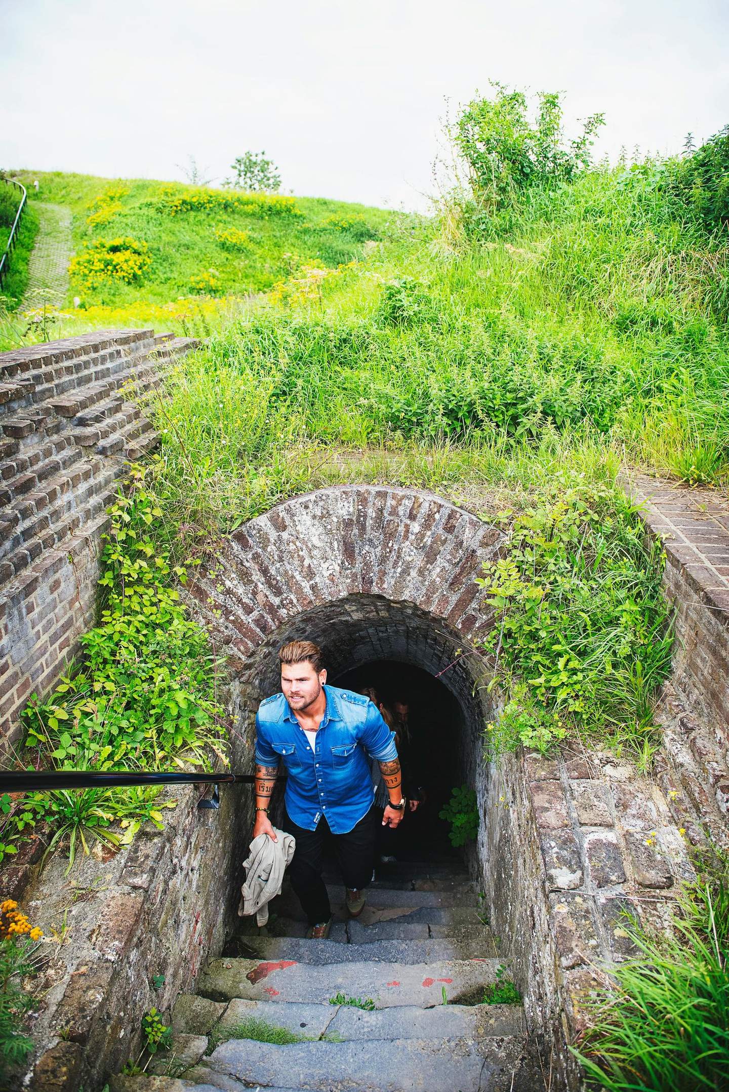 A man in a denim shirt climbs stone stairs from a brick tunnel surrounded by greenery.