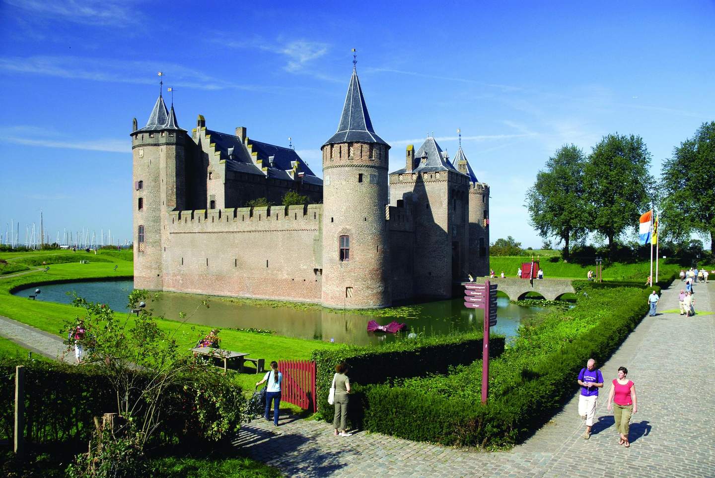 A brick castle with round towers is surrounded by a moat and green lawns; people walk near a stone path and a Dutch flag.