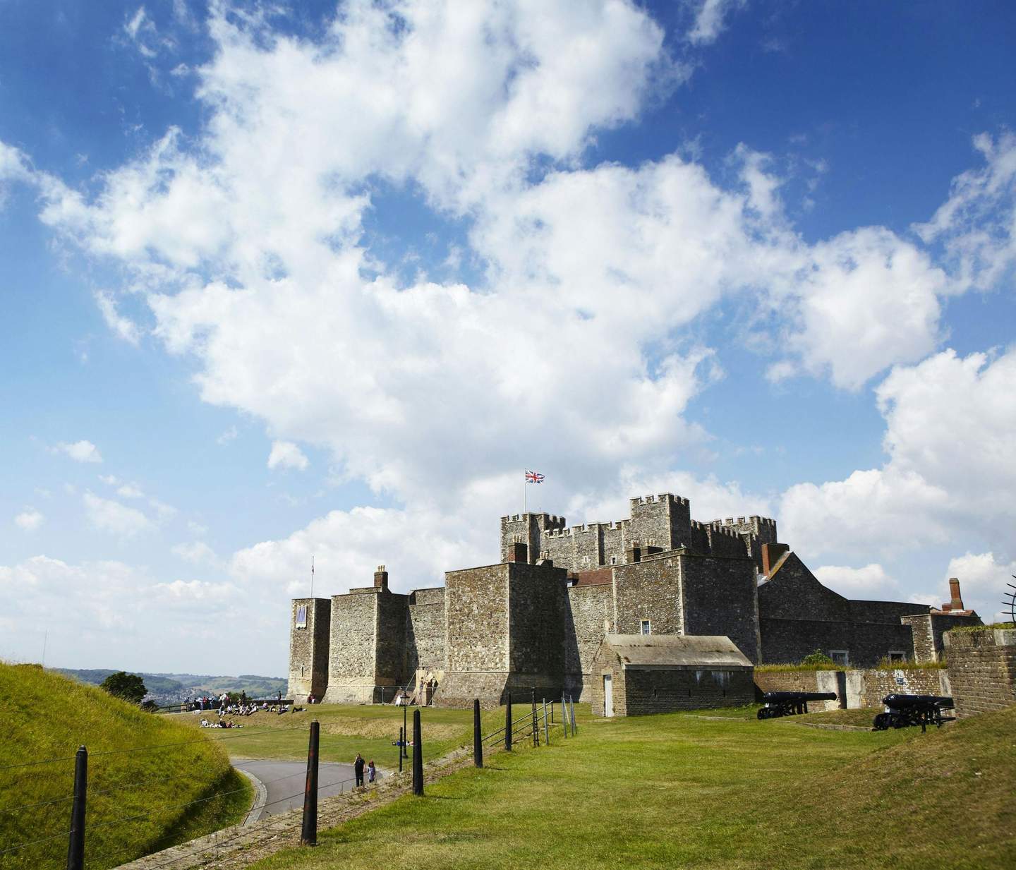 Stone castle with British flag, surrounded by grassy fields under a partly cloudy sky. Visitors walk and rest nearby.