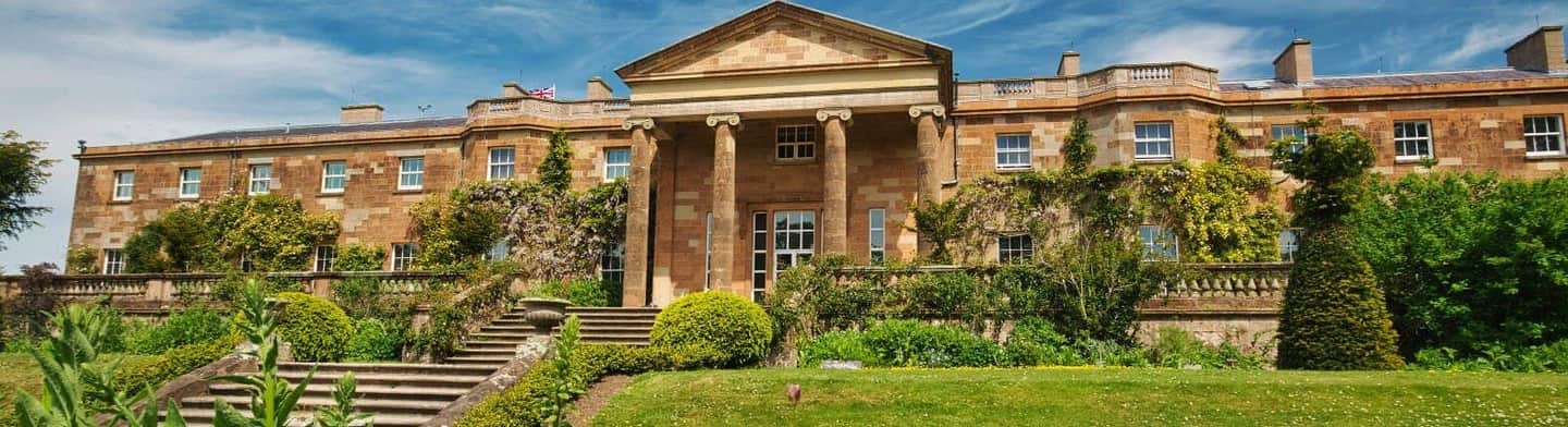 Large, historic mansion with a columned entrance, stone steps, greenery, and manicured shrubs under a blue sky.
