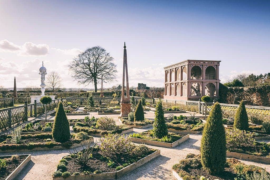 Kenilworth Castle and Elizabethan Garden A formal garden with manicured shrubs, statues, a tall obelisk, and a pink pavilion under a clear sky.
