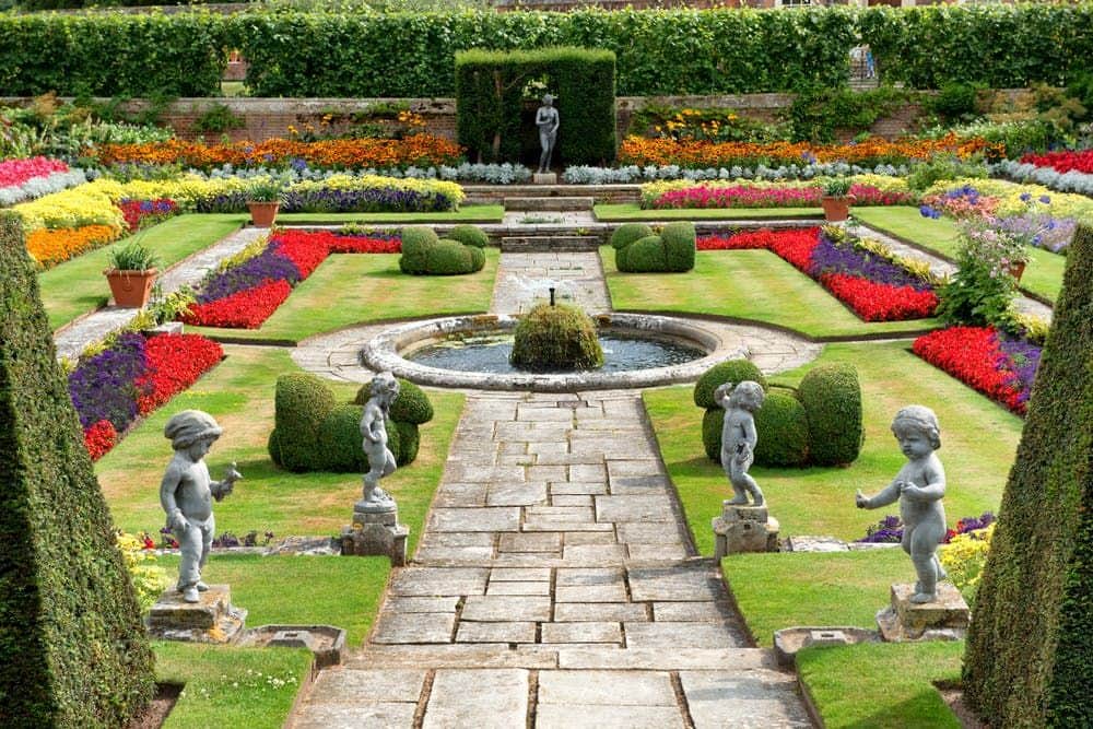 A formal garden with colorful flower beds, stone pathways, a central fountain, and statues of children positioned symmetrically.