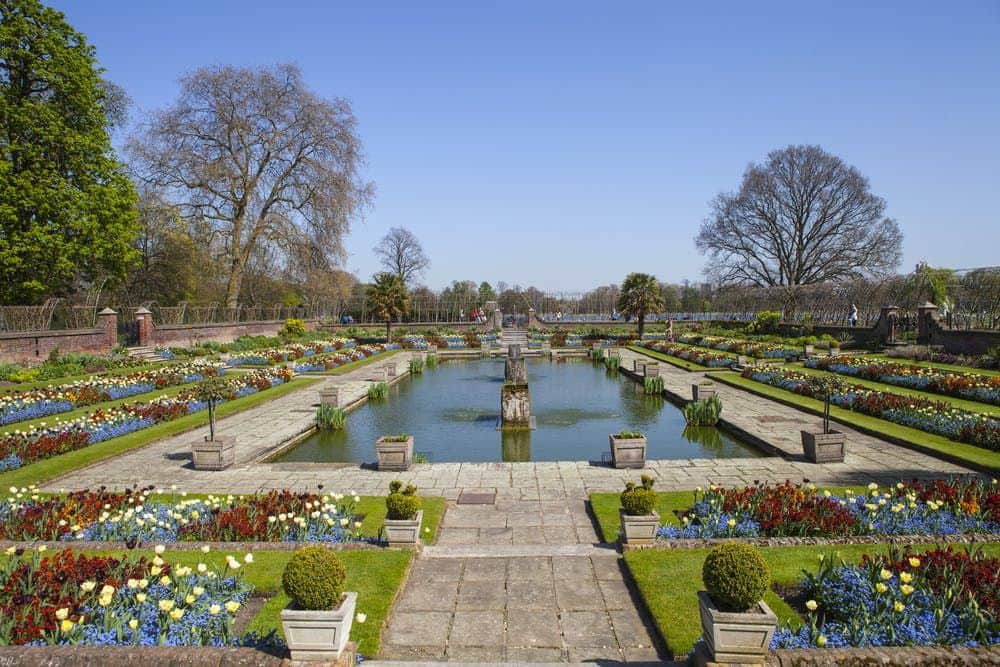 Formal garden with symmetrical flowerbeds and a central pond, bordered by pathways, trees, and manicured shrubs.
