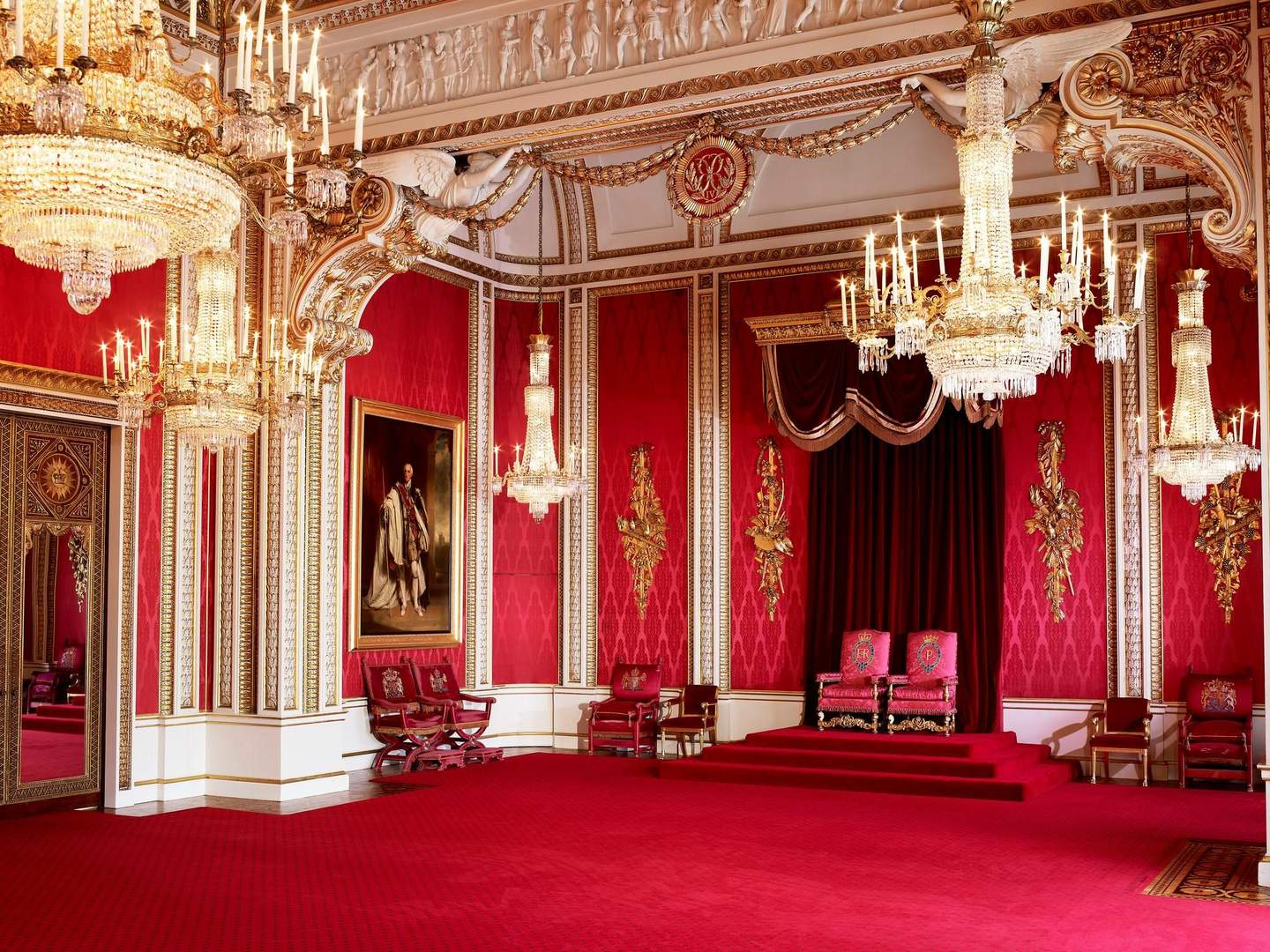Ornate room with red walls and carpet, two thrones on a dais, chandeliers, gold accents, and a portrait of a figure in royal attire.