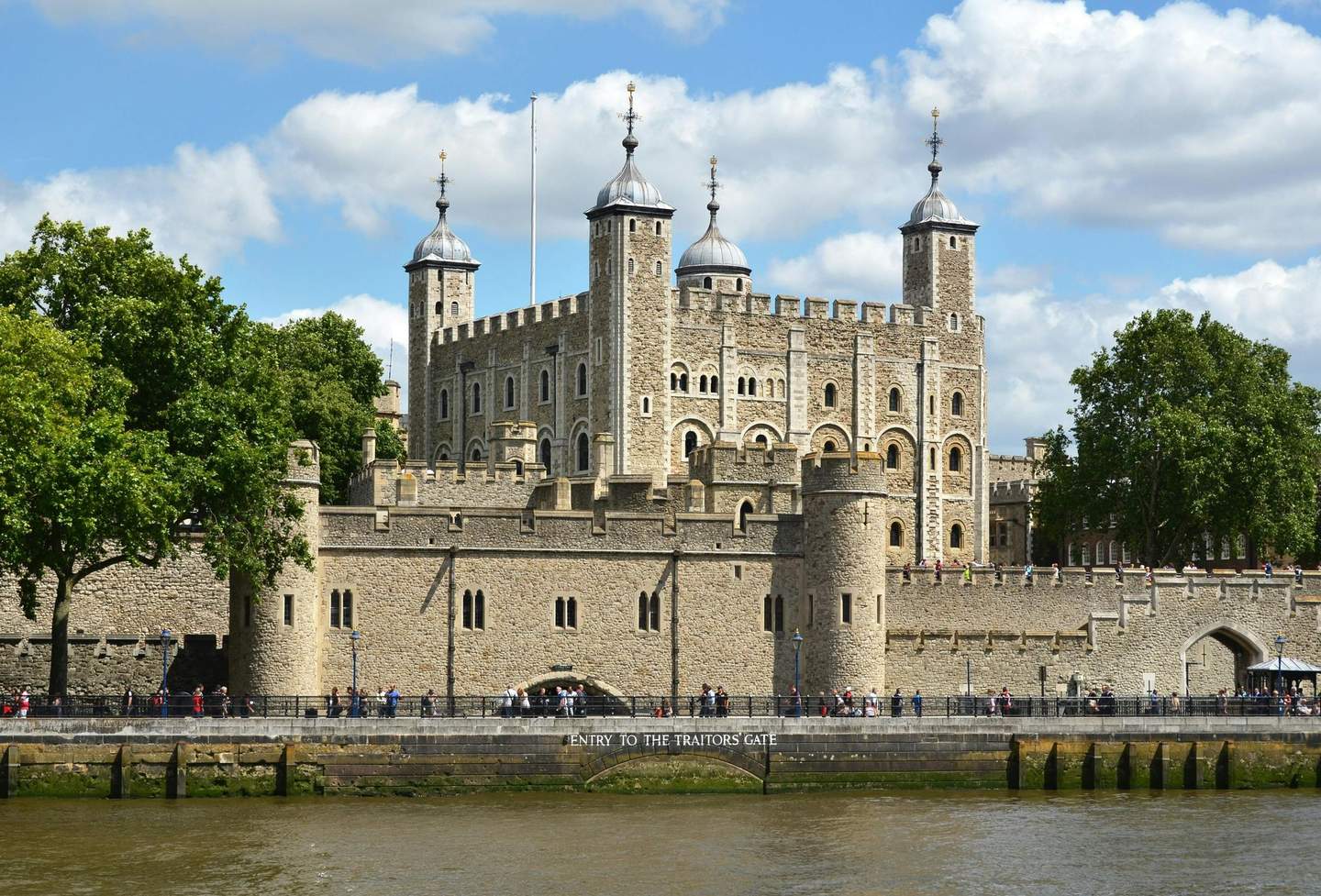 Tower of London A historic stone castle with multiple towers stands behind a river and stone wall. People are walking along the riverbank.