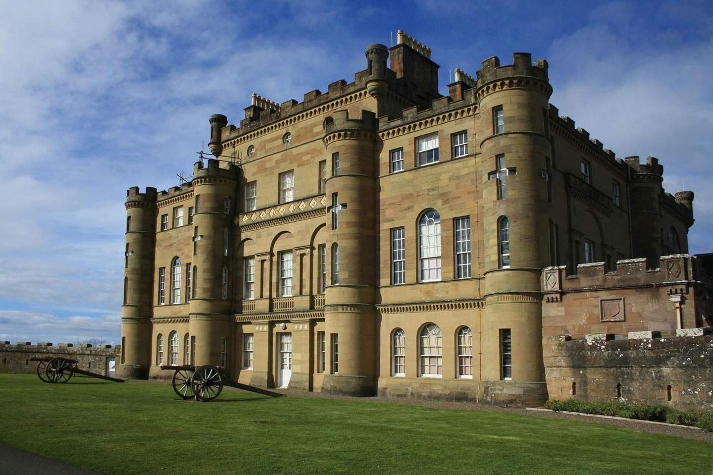 A historic castle with multiple towers and large windows, surrounded by manicured lawns, and with two cannons in front.