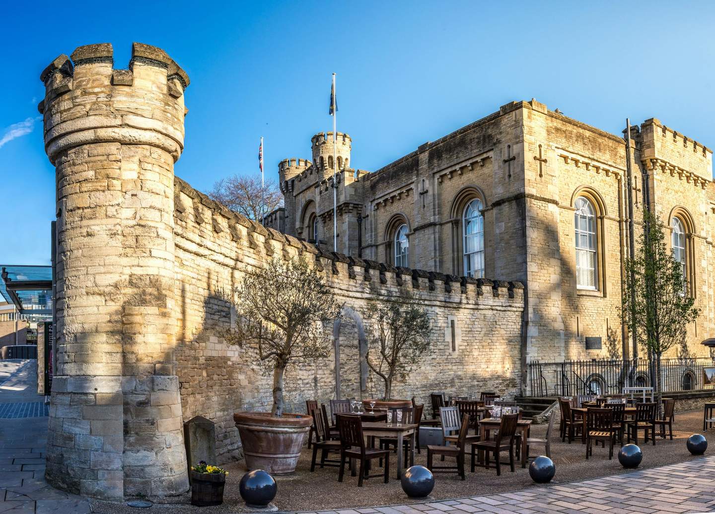 Outdoor seating area with wooden tables and chairs beside an old stone castle with crenellated walls and corner turrets.