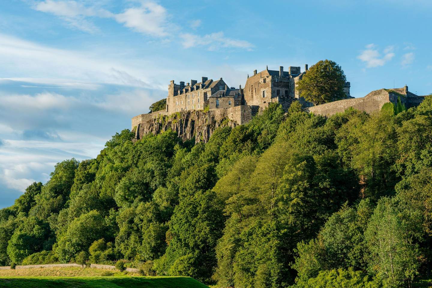 Stirling Castle Stone castle perched on a wooded hilltop under a blue sky with scattered clouds.