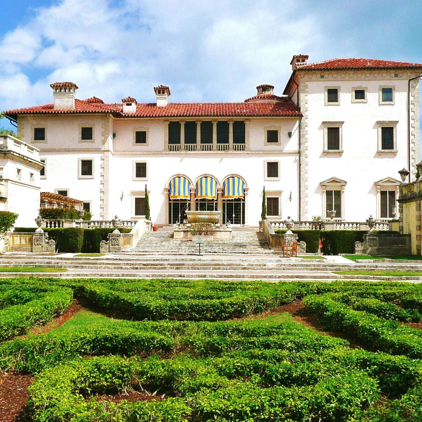 Vizcaya Museum & Gardens Elegant mansion with red-tiled roof, striped awnings, and intricate garden paths in the foreground under a partly cloudy sky.