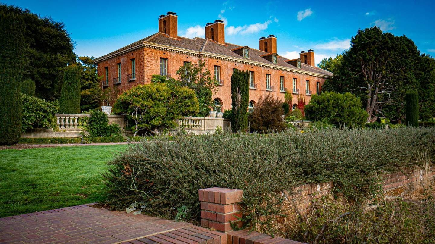 A large red-brick mansion with numerous chimneys is surrounded by lush gardens, shrubs, and a stone balustrade.