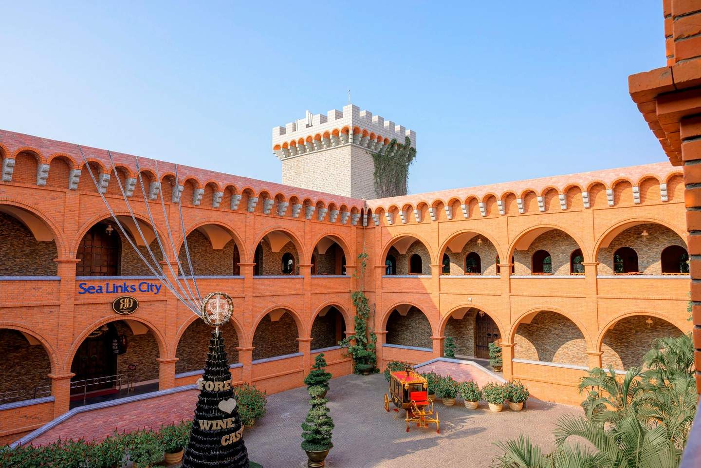 A courtyard with red brick architecture, a decorative wine display, a vintage carriage, and plants under a blue sky.