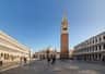 A wide plaza with a tall brick bell tower, a large ornately designed church, and people walking around on a clear day.