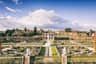 Jardin formel avec des allées symétriques, des statues, des buissons soignés et une fontaine centrale, sur fond de maisons et de ciel nuageux.